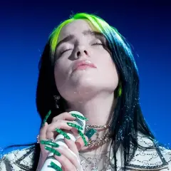 Billie Eilish z zielonymi pasemkami we włosach, z zamkniętymi oczami, śpiewa do mikrofonu. Tekst "lovely" brzmi w jej wykonaniu.