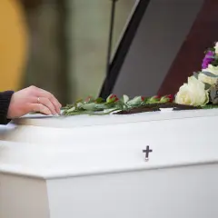 Dłoń kładzie róże na białej trumnie ozdobionej kwiatami i krzyżem.