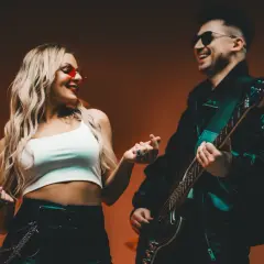Para z uśmiechem, ona w czerwonych okularach, on gra na gitarze. Ich dłonie się stykają, jakby tworzyły tekst.