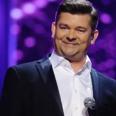 Mężczyzna w czarnym garniturze i białej koszuli, z mikrofonem. Jego uśmiech i spojrzenie podkreślają akcent przez twe oczy zielone tekst.