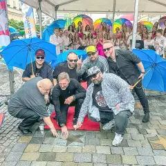 Grupa mężczyzn, prawdopodobnie zespół, odsłania gwiazdę z napisem "PIERSI BAŁKANICA" na chodniku. W tle kolorowe parasole i publiczność.
