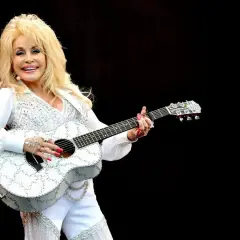 Dolly Parton, w białym, błyszczącym stroju, gra na gitarze. Wyobraź sobie, że śpiewa "Jolene", a tekst piosenki rozbrzmiewa w powietrzu.