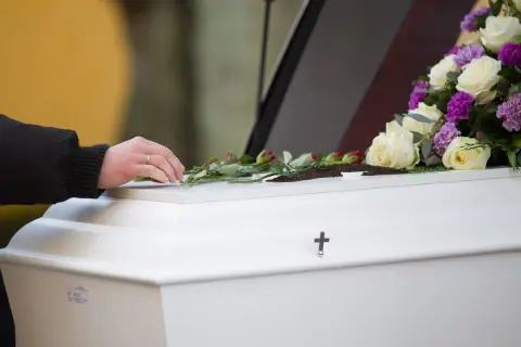 Dłoń kładzie róże na białej trumnie ozdobionej kwiatami i krzyżem.