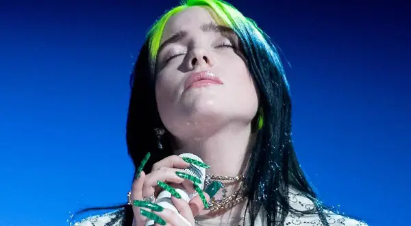 Billie Eilish z zielonymi pasemkami we włosach, z zamkniętymi oczami, śpiewa do mikrofonu. Tekst "lovely" brzmi w jej wykonaniu.