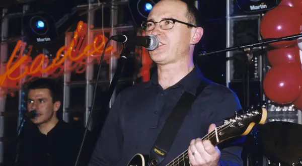 Wokalista z gitarą śpiewa na scenie, a w tle neonowy napis. Czas na koncert, a kiedy przyjdzie na ciebie czas tekst.