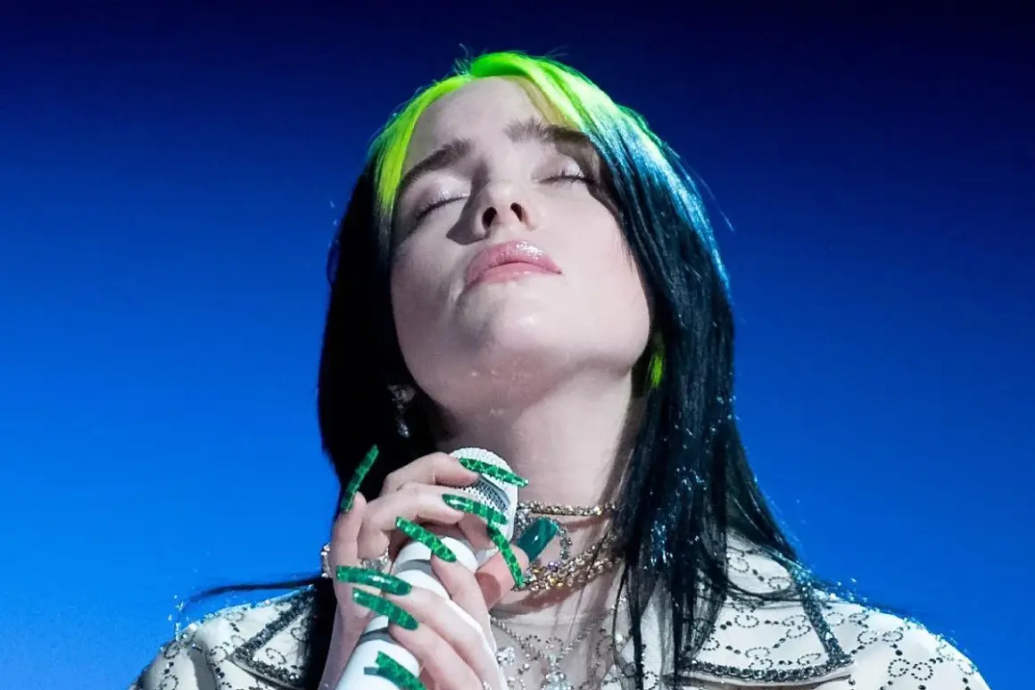 Billie Eilish z zielonymi pasemkami we włosach, z zamkniętymi oczami, śpiewa do mikrofonu. Tekst "lovely" brzmi w jej wykonaniu.