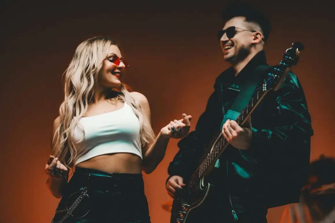 Para z uśmiechem, ona w czerwonych okularach, on gra na gitarze. Ich dłonie się stykają, jakby tworzyły tekst.