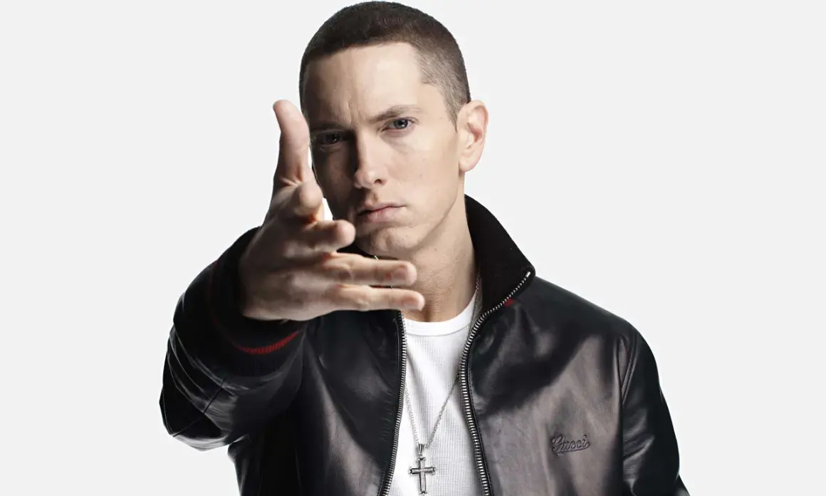 Eminem w czarnej kurtce i białej koszulce, gestykuluje ręką.