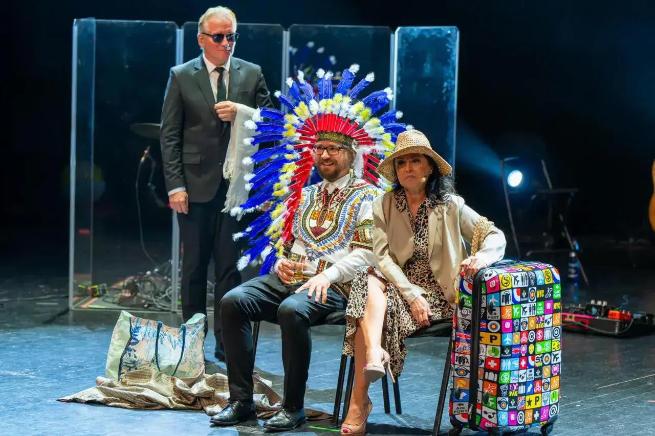 Okrutna, zła i podła scena teatralna. Mężczyzna w pióropuszu siedzi obok kobiety w kapeluszu. Stojący mężczyzna w garniturze.
