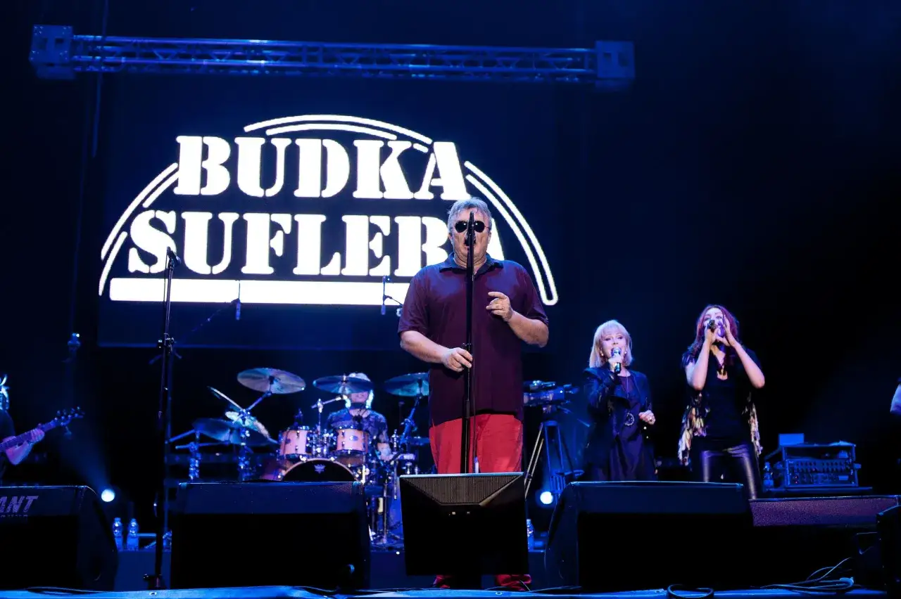Budka Suflera na scenie. Wokalista śpiewa, a za nim widać perkusistę i dwie wokalistki. Tekst