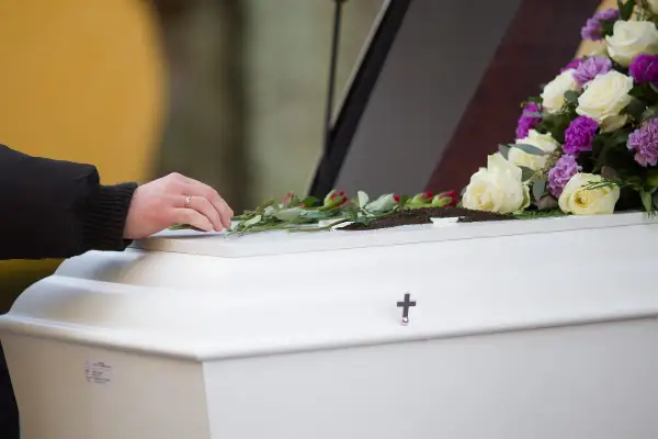 Dłoń kładzie róże na białej trumnie ozdobionej kwiatami i krzyżem.