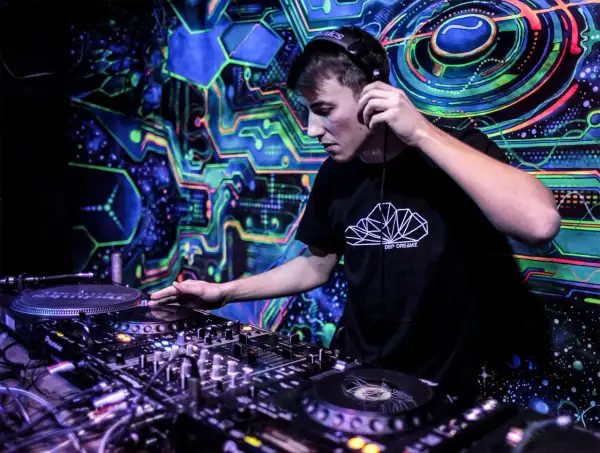 DJ w słuchawkach miksuje muzykę na tle psychodelicznej, neonowej grafiki.